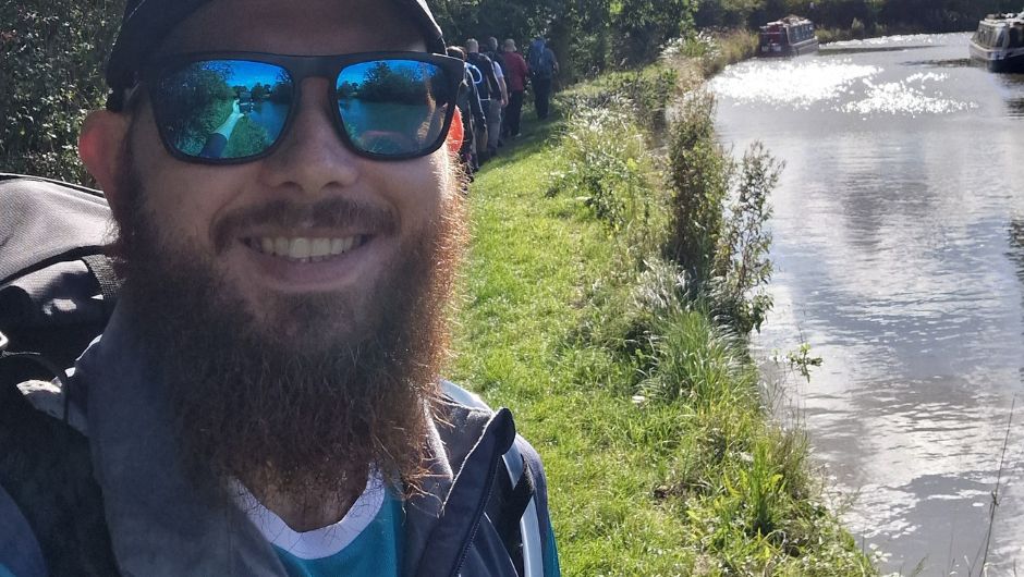 A man with sunglasses smiles at the camera, he has a large beard and is stood next to a canal. 
