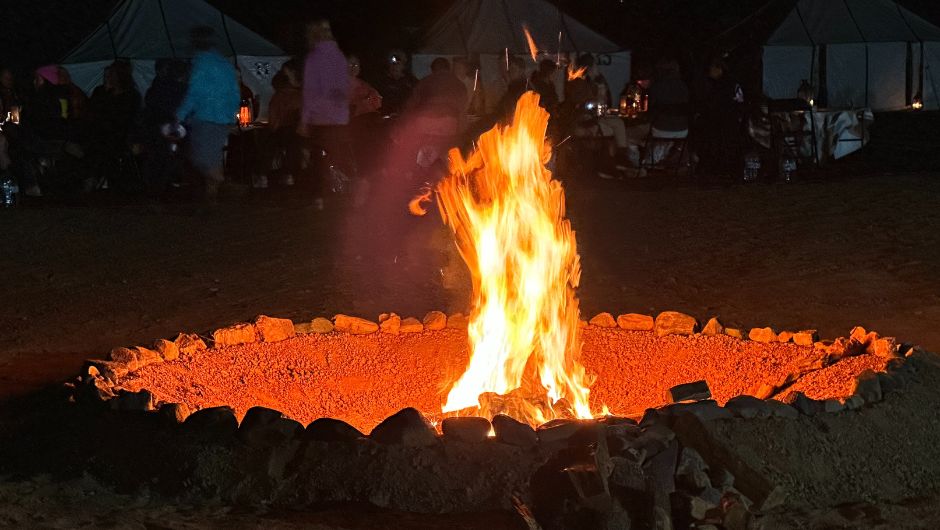 A burning bonfire in a fire pit sits at the front of the image with blurred people in the background. 