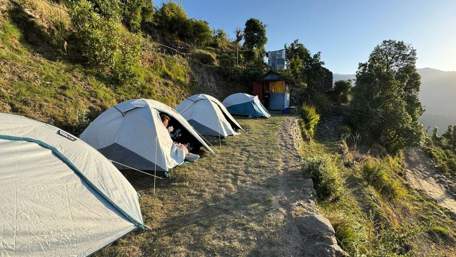 a campsite of pitched tents, showing a woman sat looking out of her tent enjoying a hot drink as the sun sets