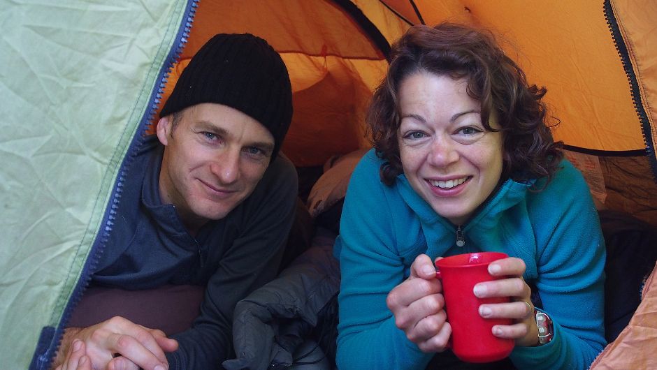 two people smiling inside their tent, keeping warm with a hot drink
