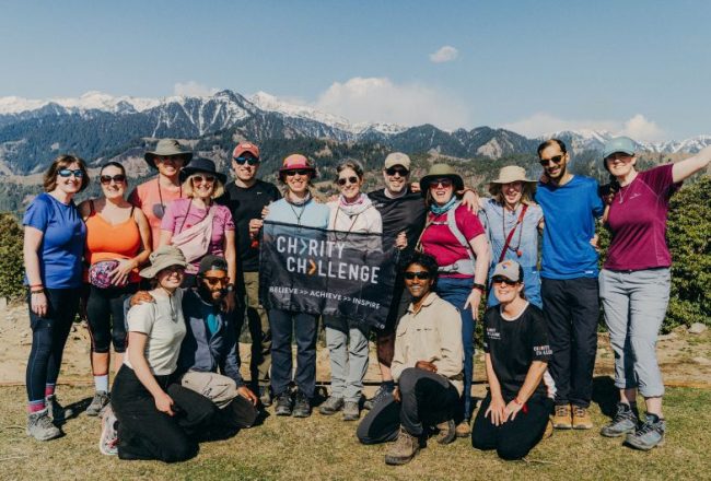 a group of smiling challengers posing with a Charity Challenge flag, surrounded by Indian Himalaya mountains