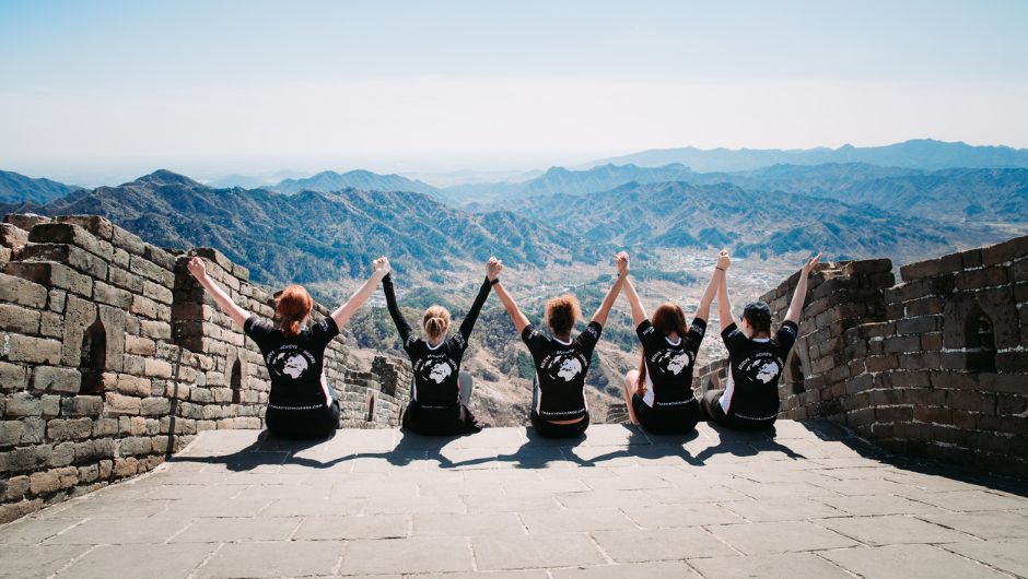 5 people sitting in a row, on the edge of some steps on the Great Wall of China. They are all holding their hands in the air, celebrating and looking out at the view.