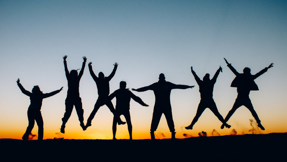 A group of silhouetted people jumping in celebration against a warm orange sunset and blue sky.