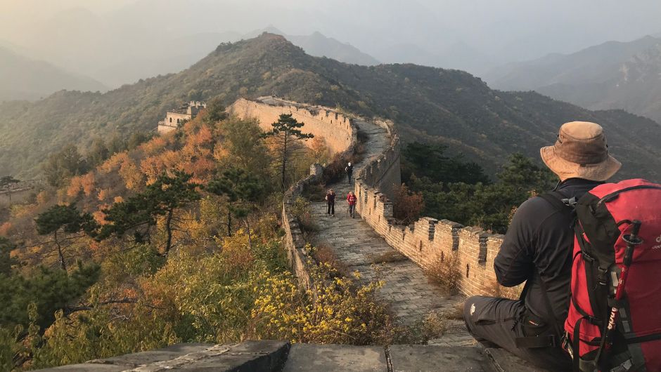 View of a section of the Great Wall of China, with mountains in the background and a singular trekker sitting in the foreground looking at the view.
