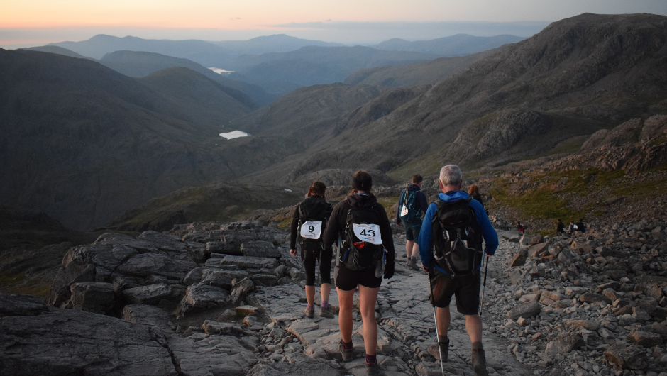 A group of people trekking down a UK mountain, as the sun rises in the distance.