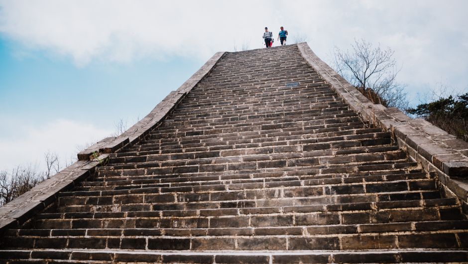 View from the bottom of a set of steep stairs on a section of the Great Wall of China, with two trekkers at the top about to descend. 