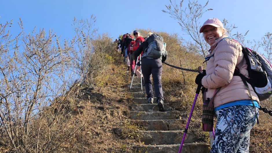 A group of smiling trekkers, climbing up some steep stairs of the Great Wall of China with walking poles.