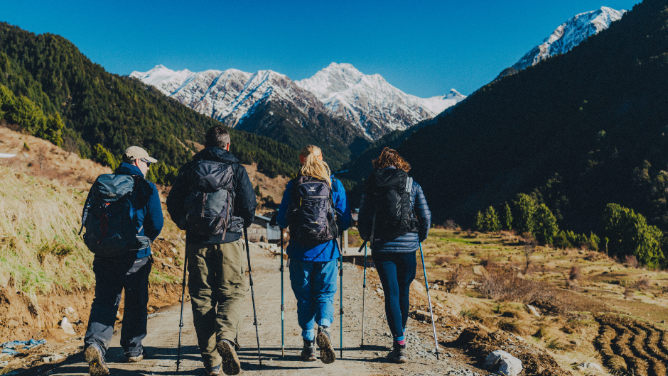 Four people dressed in hiking gear, walking down a path leading towards the Himalayan Mountains in India.