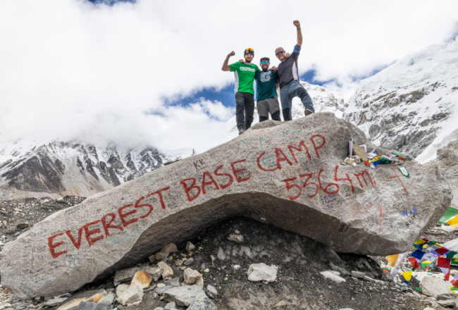 Three people stood cheering on a rock that's signed Everest Base Camp