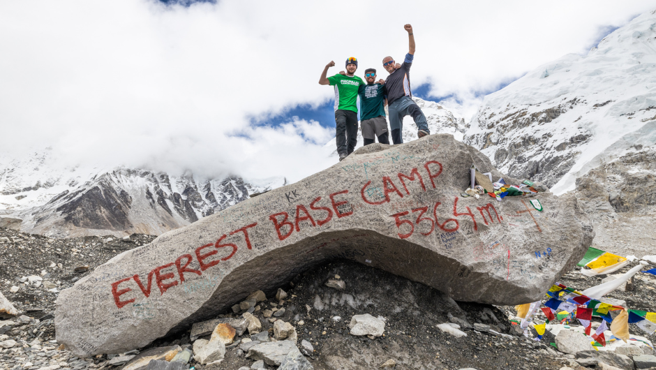 Three people stood cheering on a rock, signed with Everest Base Camp, surrounded by snowy mountains.