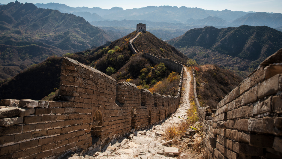 A section of the Great Wall of China. showing worn, uneven steps, the walls and a turret in the background, set against surrounding rolling mountains.