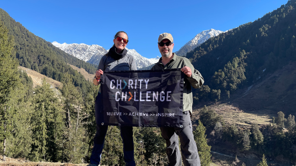 Two trekkers holding a flag that reads Charity Challenge Believe Achieve Inspire, in front of mountainous Himalayan scenery, trees and a clear blue sky.