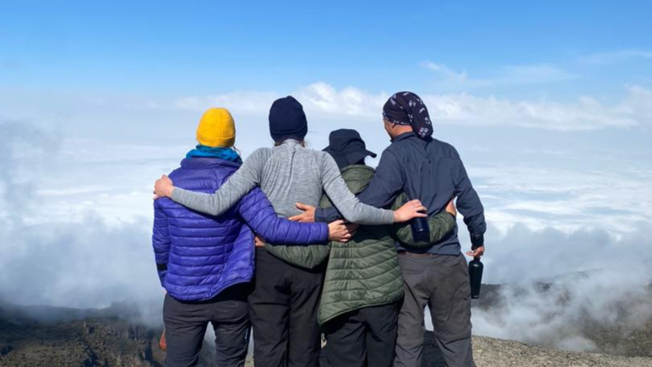 Four Mount Kilimanjaro trekkers, with their arms around each other, looking out at the clouds from the summit.