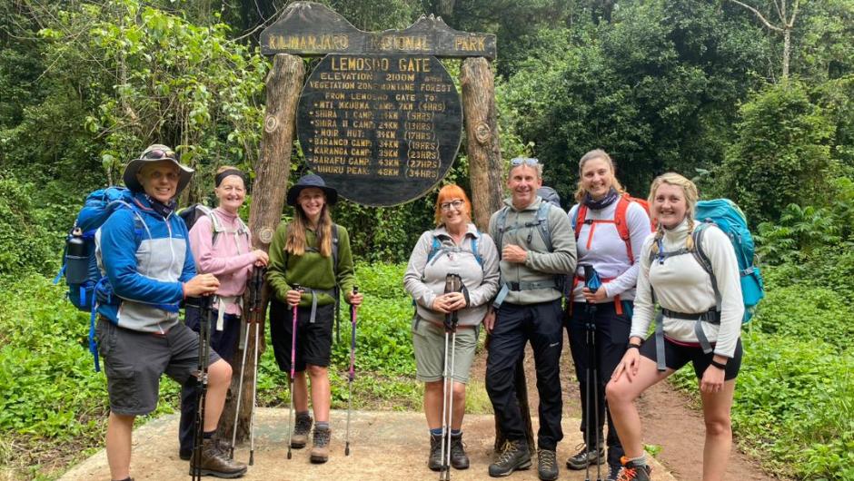 A group of hikers smiling in front of the Kilimanjaro National Park, Lemosho Gate sign, in Tanzania, Africa.