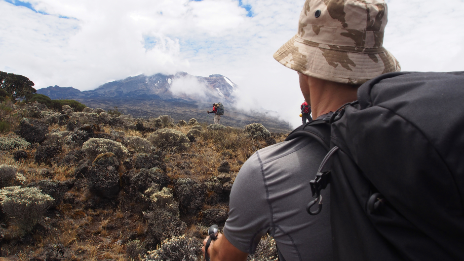 A distance view of Mount Kilimanjaro, as a man dressed in hiking gear sips water from his camel pack during a trek.