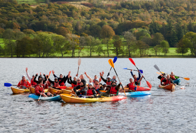 A group of people in kayaks in the centre of Coniston Water in the Lake District, with their paddles lifted in the air.