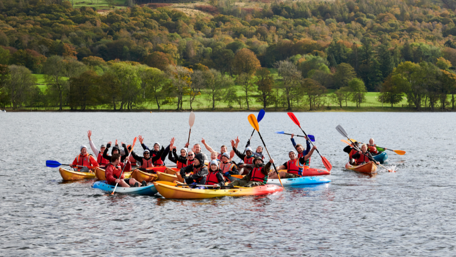 A group of people in kayaks in the centre of Coniston Water in the Lake District, with their paddles lifted in the air.