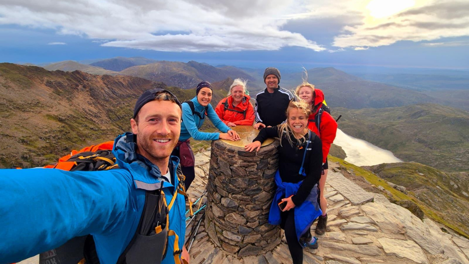 A group of smiling people posing for a selfie at the trig point on the peak of a mountain during the National Three Peaks Challenge.