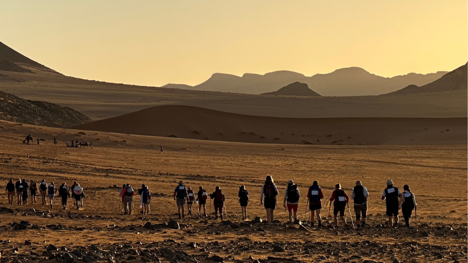 A line of trekkers in the Sahara Desert, walking across sandy desert plains under an orange sky.