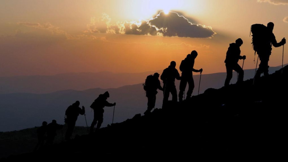 A silhouetted line of people hiking up a steep hill, backlit by an orange sunrise