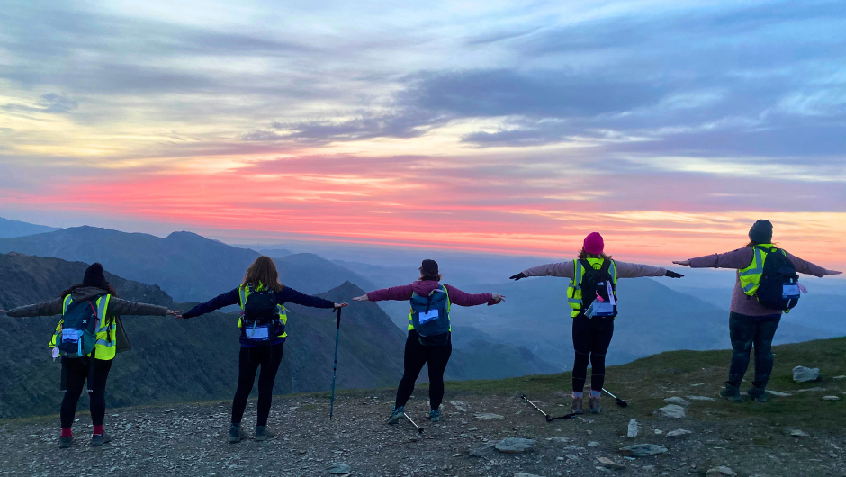 5 trekkers stood in a line on Snowdon in Wales, with their arms stretched out, in front of a pink sunrise skyline.
