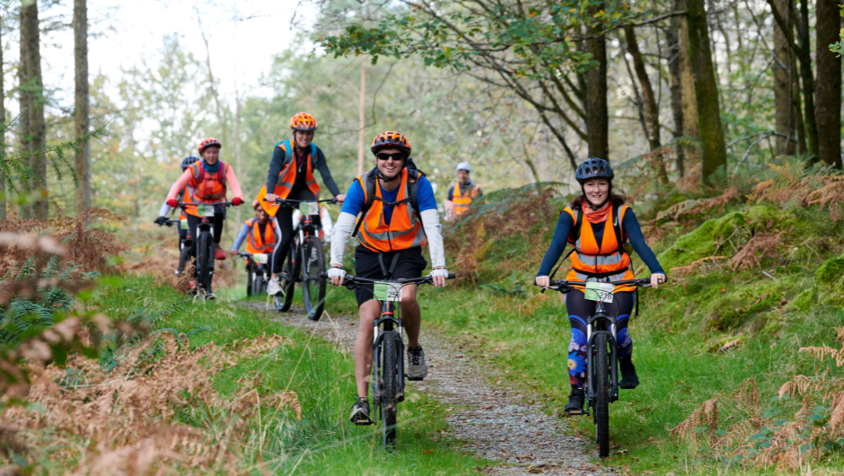 A group of smiling people riding bikes through a section of Grizedale forest in the Lake District.