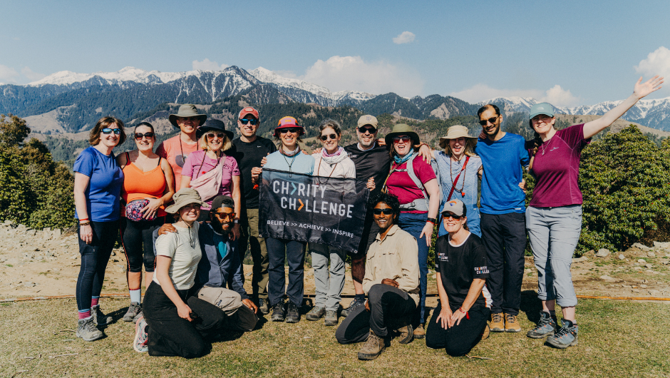 A group of trekkers posing in front of a section of the Himalaya in India, with a Charity Challenge flag being held up in the middle of the group.