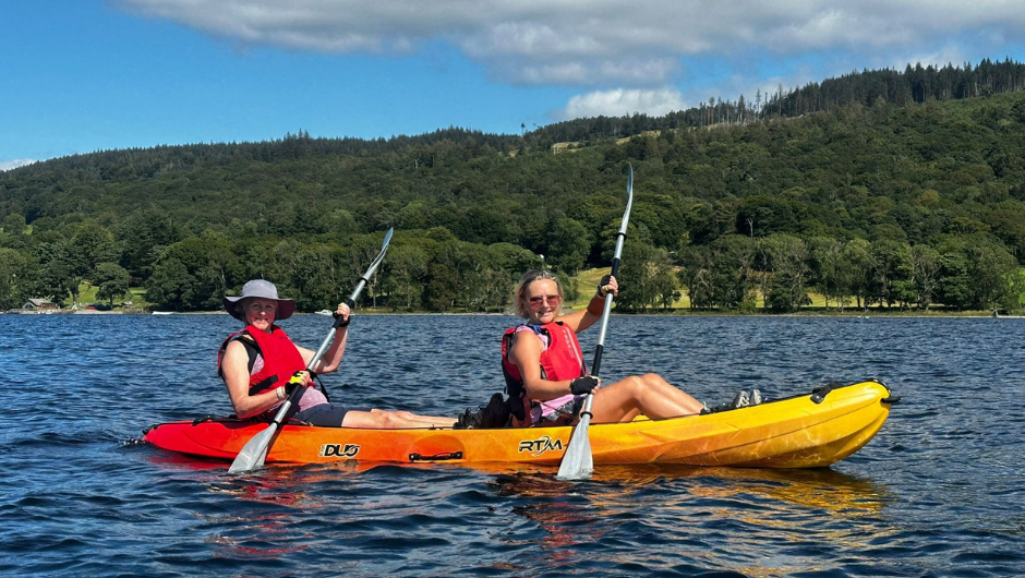 Two people in a two-person kayak, smiling as they paddle across Coniston Water during the Lake District Triple Challenge.