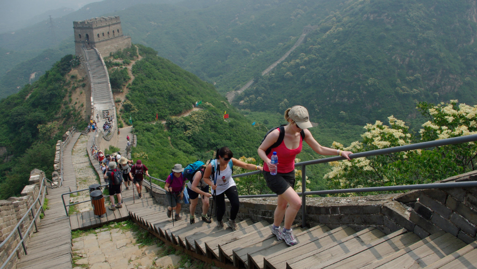 Trekkers climbing some steps on a section of the Great Wall of China, with a watchtower in the background, and lush greenery surrounding the wall.