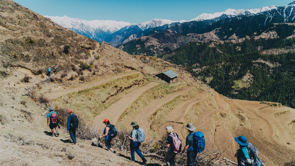 A group of trekkers, walking alongside some rural farmland in the Himalayas, with some snow-capped peaks of the Himalaya mountains in the background.