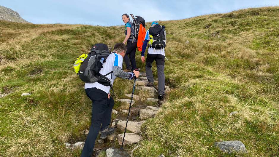 Three trekkers climbing some rocky steps on a section of a hike up to the summit of Old Man of Coniston in the Lake District.