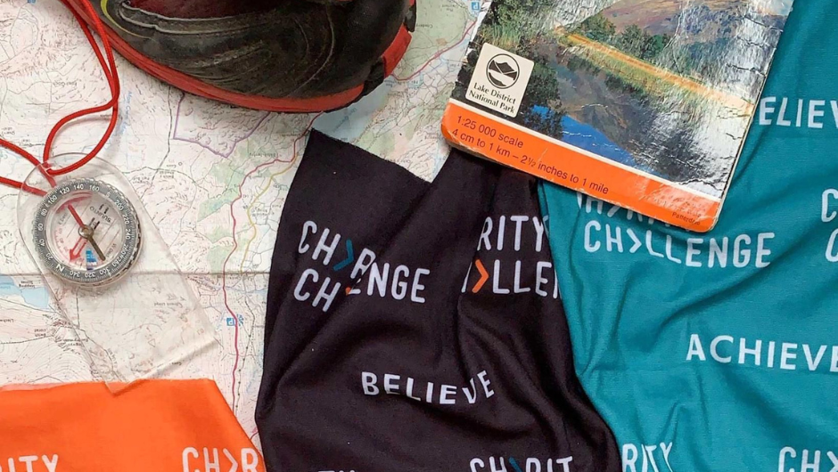 an orange, black and teal Charity Challenge neck buff laid out over a map, along with some walking boots and a compass