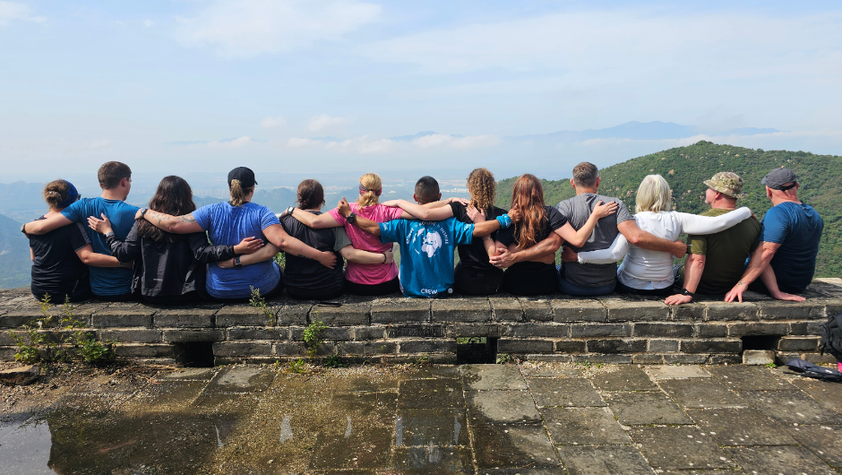 A line of people sitting on a section of the great wall of china with their arms around each others shoulders, looking out at the Yan mountain rage.