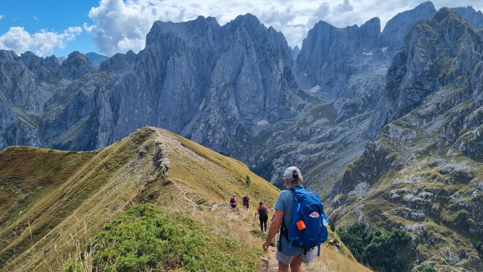 Trekkers crossing the high ridge of a grass-covered mountain in the Albanian Alps, surrounded by a contrasting wall of grey, jagged mountains.