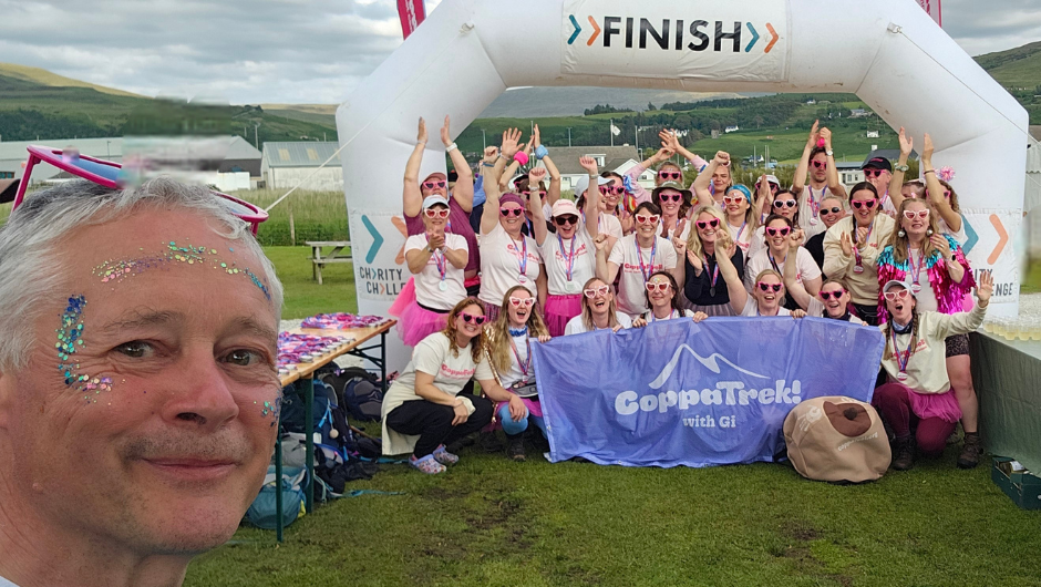 A man wearing glitter on his face smiling in front of a group of trekkers at a finish line. They are all smiling and celebrating, holding up a CoppaTrek! with Gi flag.