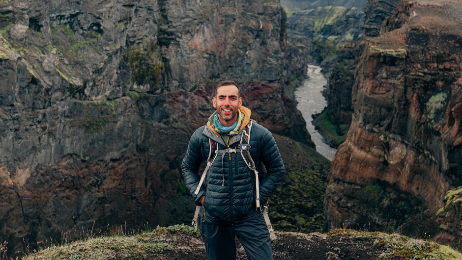 A man dressed in hiking gear standing and smiling in front of a view of rocky cliffs and a distant stream on the Isle of Skye.