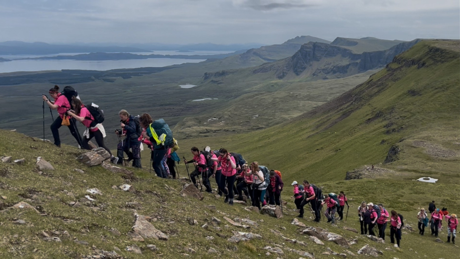 A line of people trekking up a steep in on the Isle of Skye. The background shows the rugged terrain of the Isle of Skye a distant body of water.