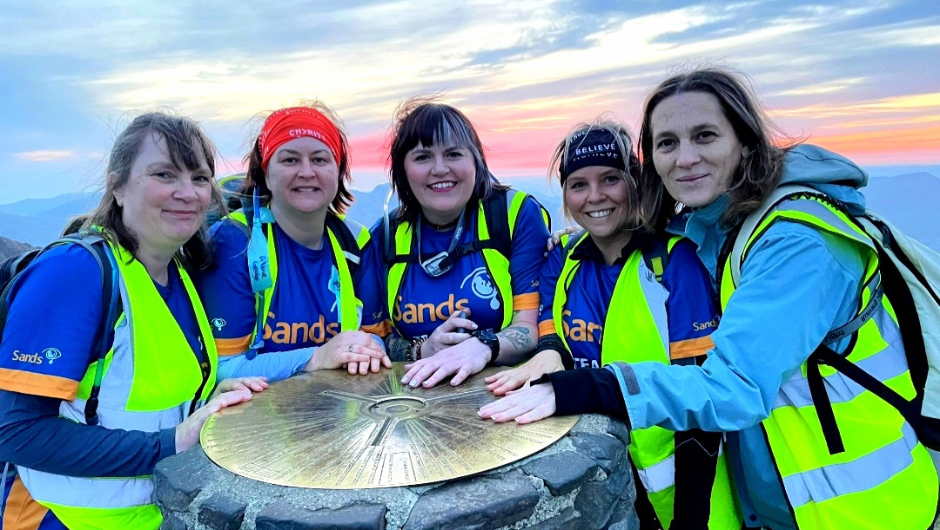 5 trekkers wearing Sands Charity tshirts placing their hands on the trig point at the summit of Snowdon (Yr Wyddfa).