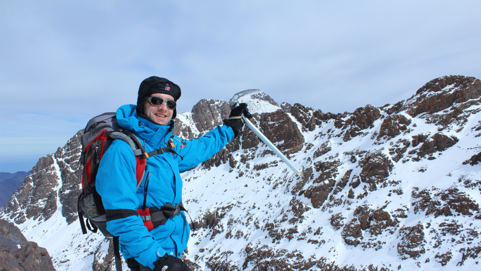 A trekker dressed in cold hiking gear, smiling as he holds up his ice pick on a section of the Mount Toubkal Trek in Morocco.