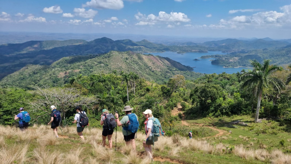 A line of trekkers travelling across a large hill in Cuba, against a backdrop of green palm trees, lush forests, bright blue water and small islands.