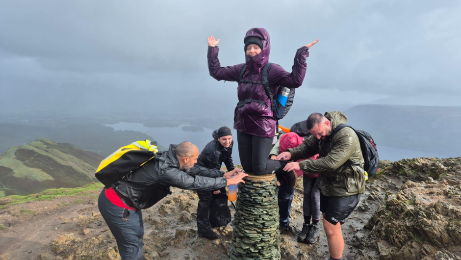 A group of trekkers at the summit of a UK mountain. A person is kneeling on the top of a trig point, whilst the people around her bow down in a funny celebration post pose.