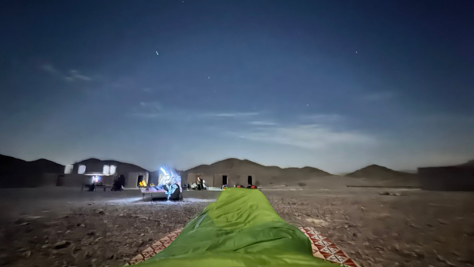 A nighttime view from inside a sleeping bag laid out on the desert ground in Jordan, facing a small camp with tents and people illuminated by soft lights. Low hills sit in the distance beneath a starry sky.