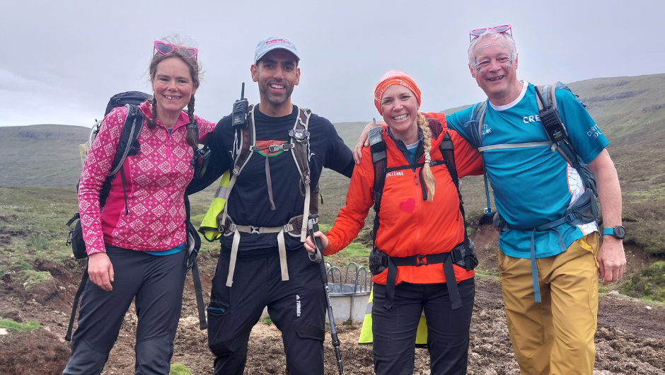Four mountain leaders smiling together, arm in arm, surrounded by some green rolling hills during a UK trek.