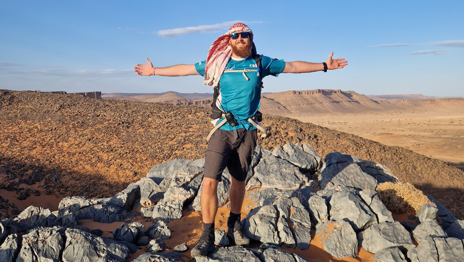 A trekker standing on a rocky section of the Sahara Desert, smiling with his arms spread directly outward.