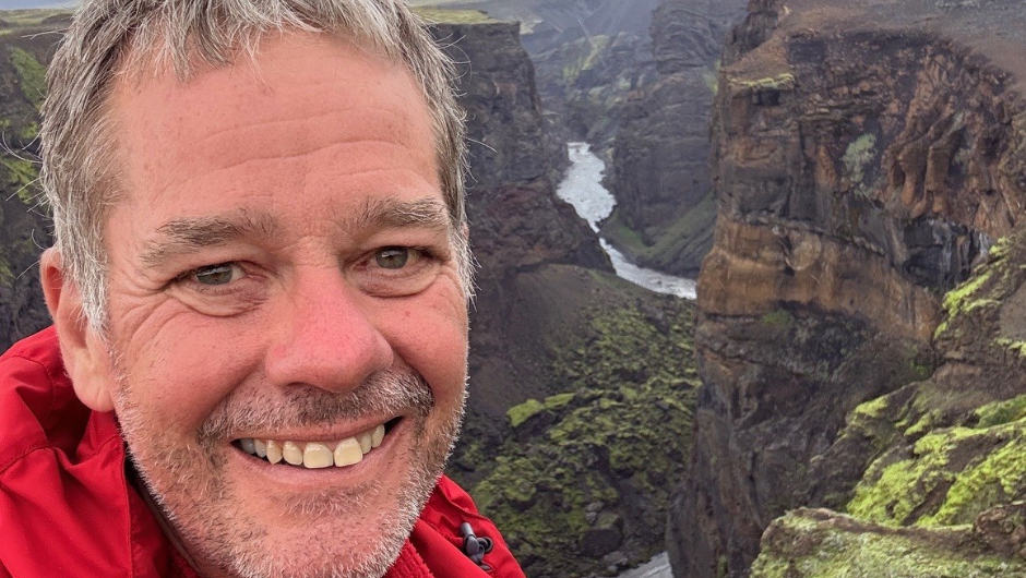 A man smiling for a selfie in the Isle of Skye, in the background are rocky cliffs and a distant river.