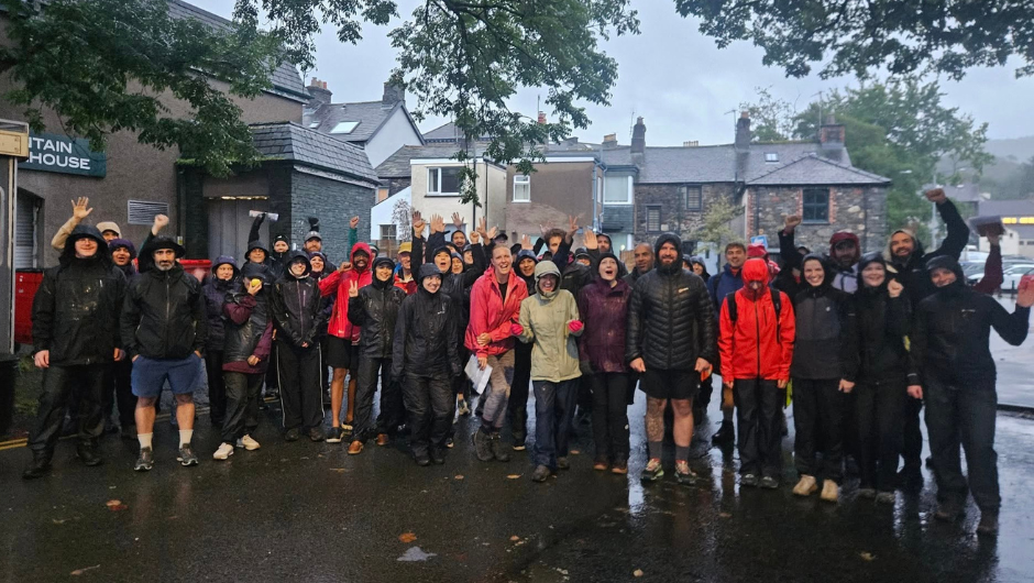 A large group of people cheering together in the rain, dressed in rainy hiking gear during The Race The Lakes Challenge with Capital One UK, in aid of BRACE Dementia Research Charity.