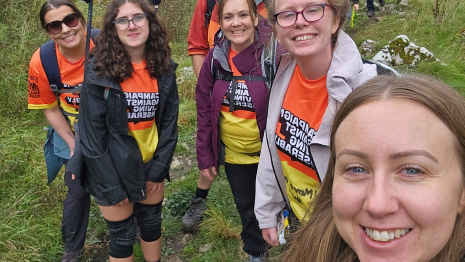 A smiling woman taking a selfie with some trekkers in the background also smiling.