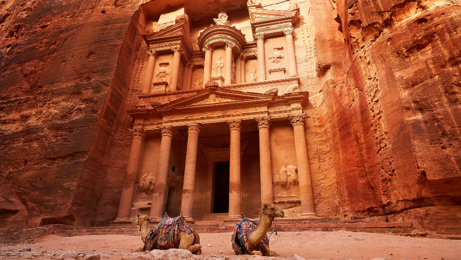 The iconic Treasury of Petra in Jordan, carved into rose-red rock cliffs, with two camels resting in the foreground. A stunning view of this ancient archaeological wonder.