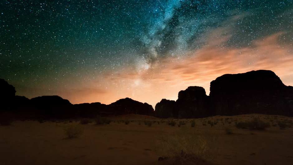 A night sky over Wadi Rum, with the Milky Way glowing above rugged desert rock formations and soft light on the horizon.