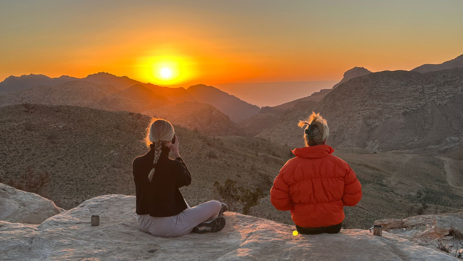 Two trekkers sat on a cliff edge, overlooking the rocky mountains of Jordan, as a glowing orange sun sets in the background.